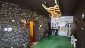 Ein Zimmer mit grünem Teppich, einer Tafelwand mit verschiedenen Aufschriften, einem kleinen Tisch und einem Ventilator.