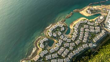 An aerial view of a coastal resort with a crescent-shaped layout, featuring a mix of villas and modern buildings, surrounded by clear blue waters and a lush green landscape.