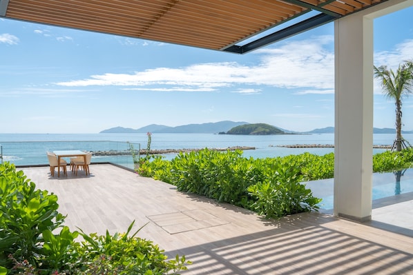 A modern outdoor patio with a wooden table and chairs, surrounded by lush greenery and overlooking a clear blue sea with distant islands. - Villa Le Corail, A Gran Melia Hotel (Nha Trang)