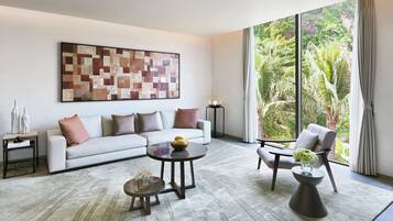 Una sala de estar moderna con un sofá, dos sillones, una mesa de centro y una lámpara de pie. Un cuadro abstracto de gran tamaño decora la pared, y una ventana ofrece vistas a exuberante follaje.