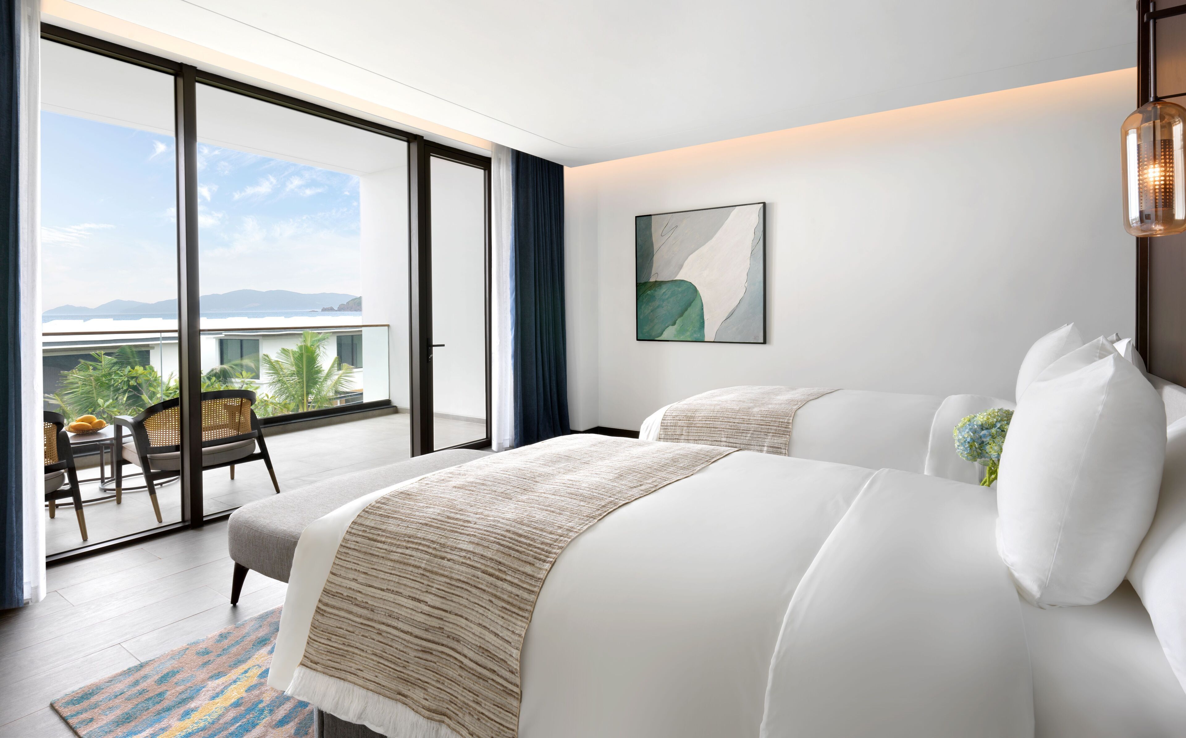 A modern hotel room with a balcony, a bed with white linens, a large abstract painting, and a hanging light fixture.