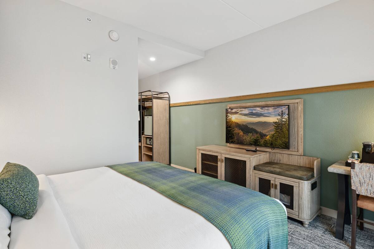 A hotel room with a bed, a desk, a chair, a painting on the wall, and a small cabinet.