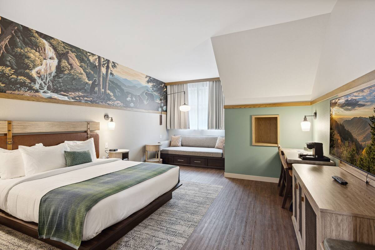 A hotel room with a large bed, a desk with a chair, a small table, and a painting on the wall.