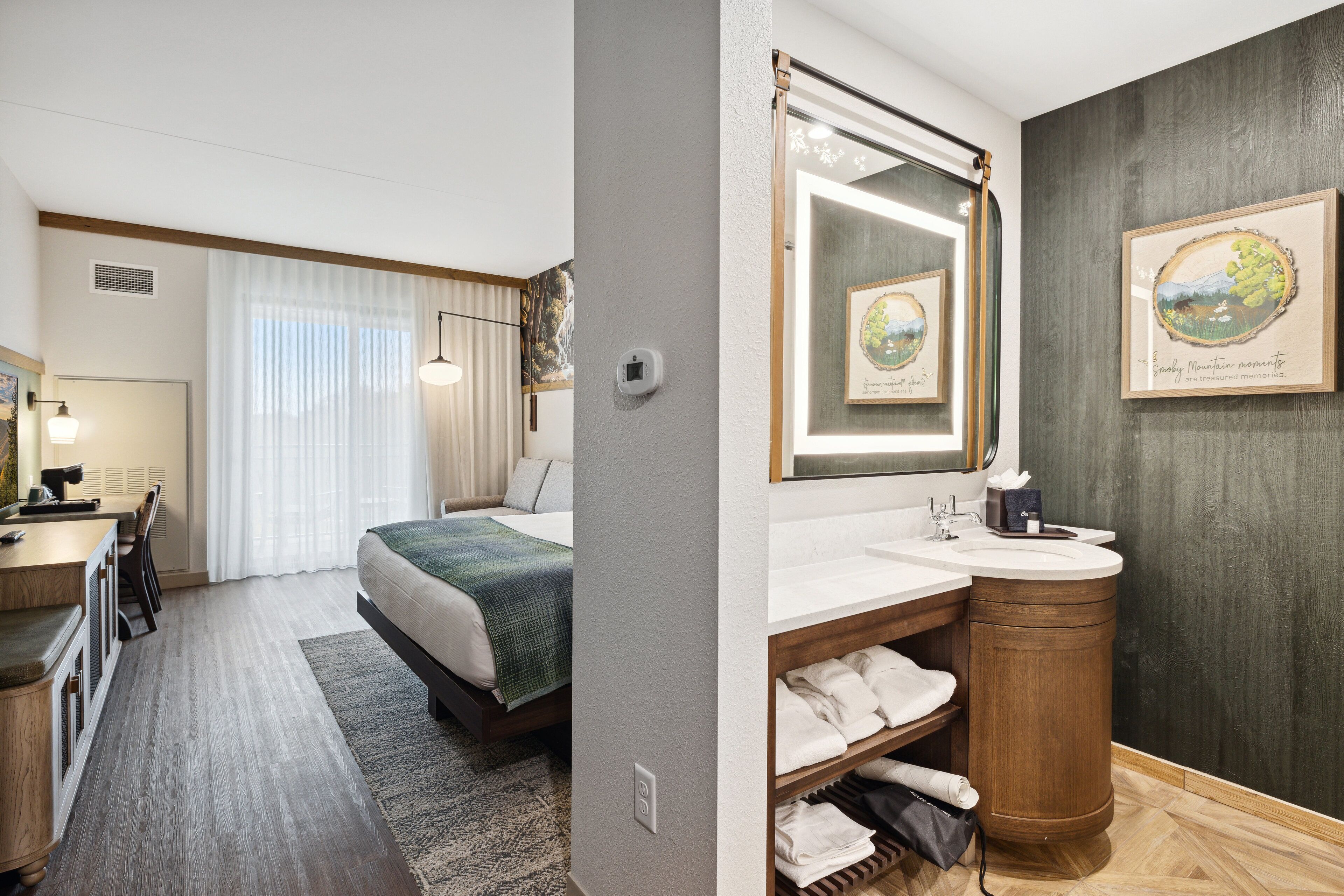 A hotel room with a bed, a desk, a bathroom with a sink and mirror, and a framed picture on the wall.
