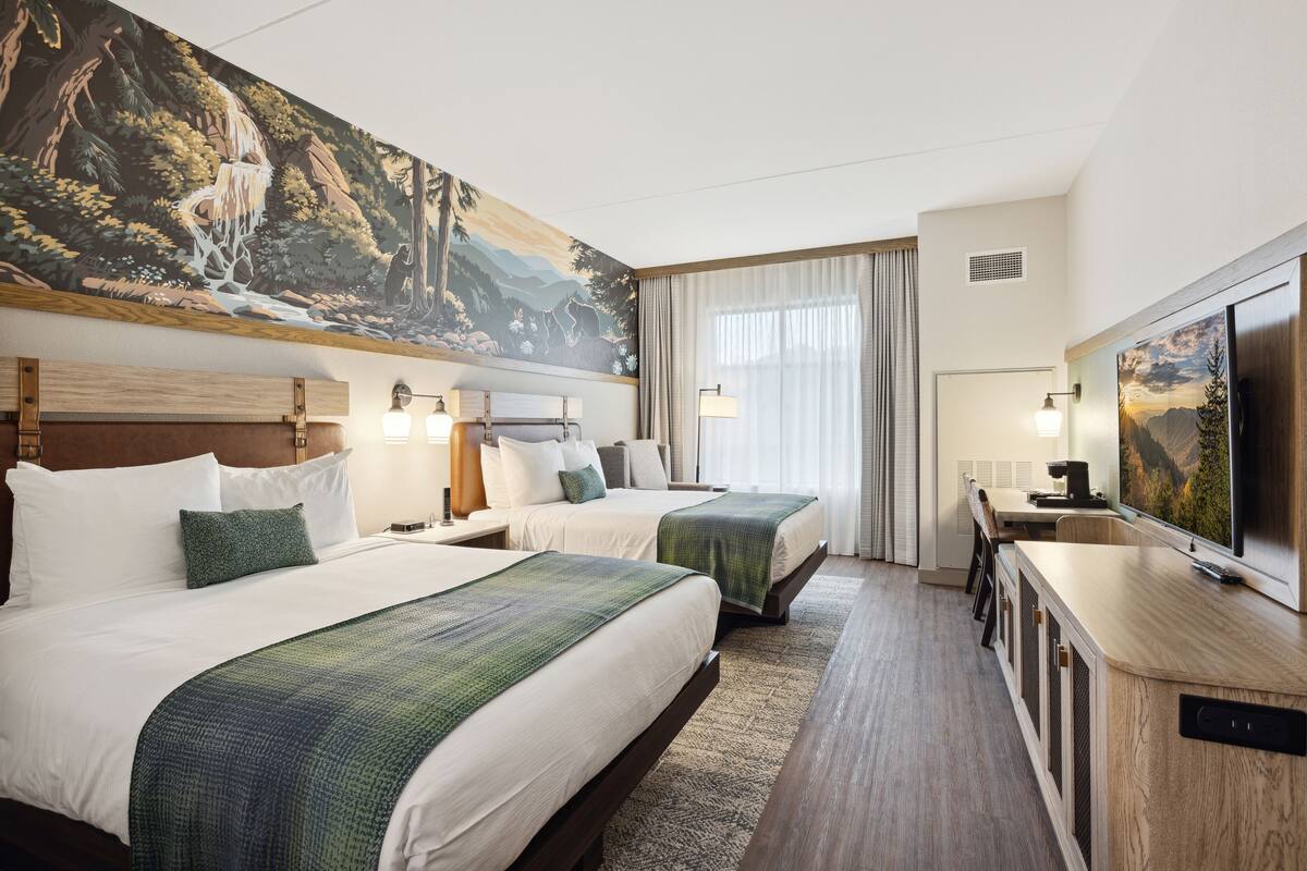 A hotel room with two beds, a desk, a TV, and a large painting of a waterfall.