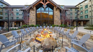 A fire pit surrounded by Adirondack chairs in front of a building with a stone facade and large windows. - HeartSong Lodge & Resort (Pigeon Forge)