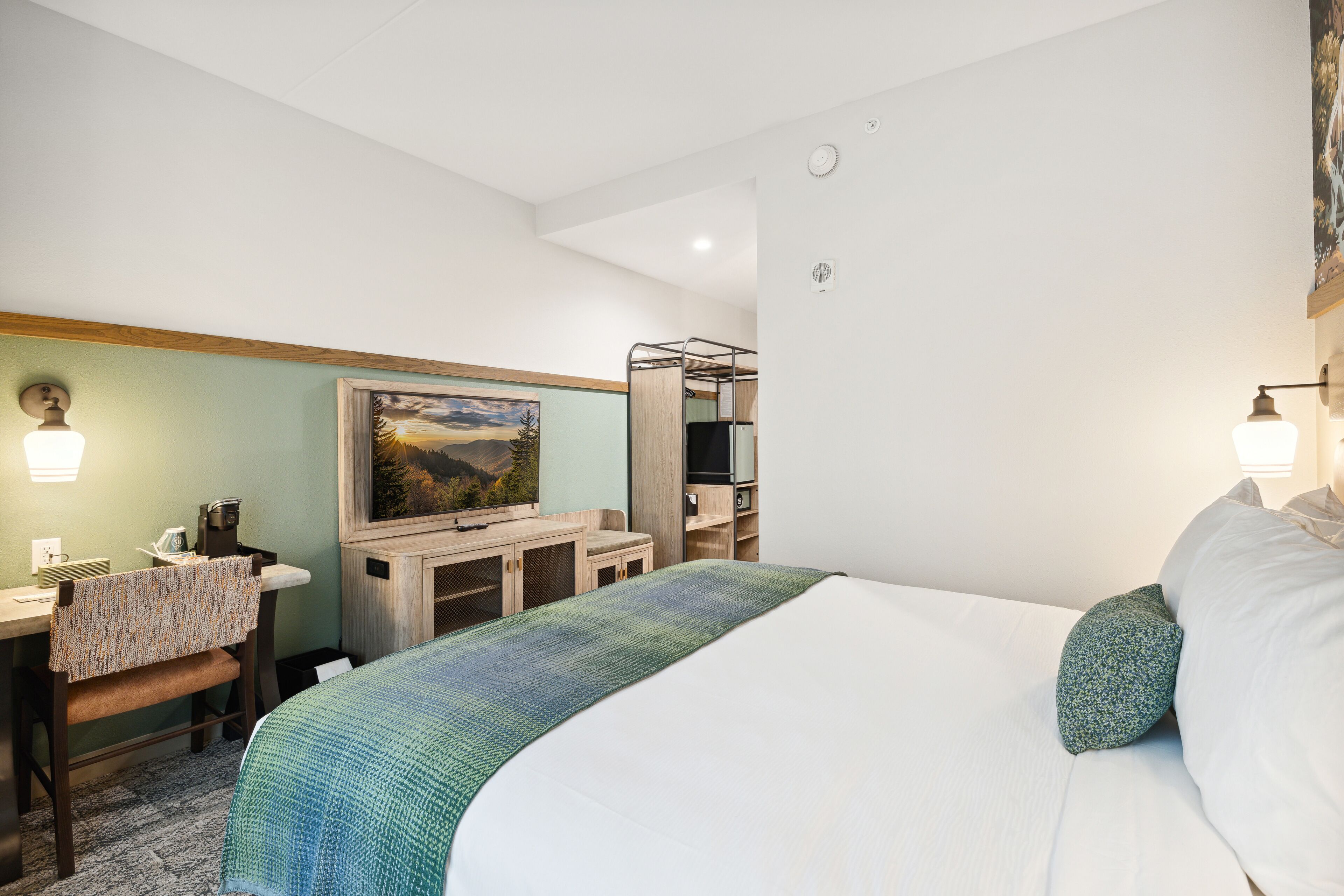 A hotel room with a bed, a desk with a chair, a television, and a painting on the wall.