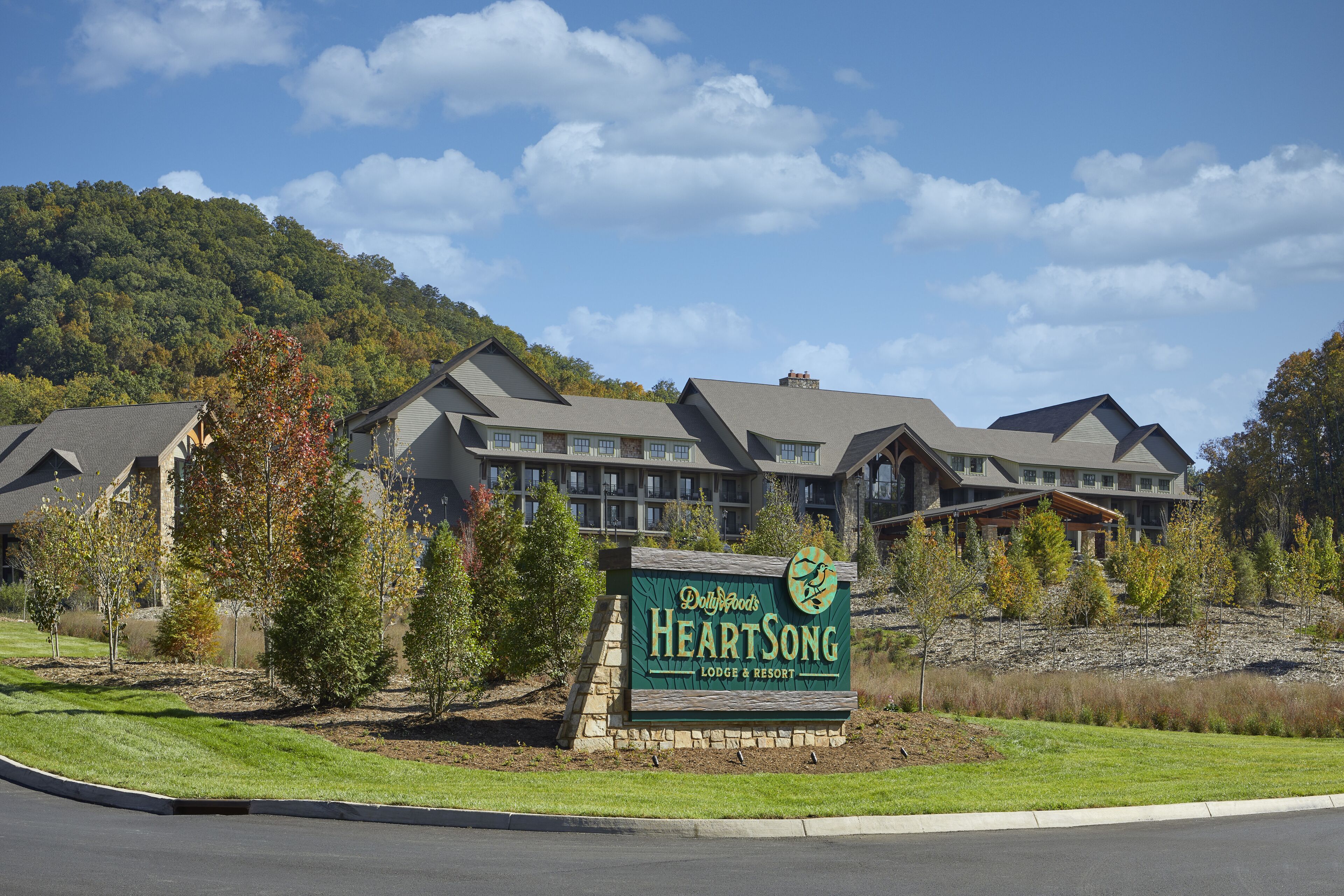 Uma grande hospedaria com vários edifícios, uma placa com a inscrição 'Dollywoods Heartsong Lodge & Resort' e um cenário de colinas ao fundo.