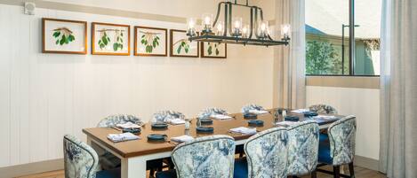 Un comedor con una mesa larga, sillas tapizadas en azul, adornos vegetales enmarcados y una araña.