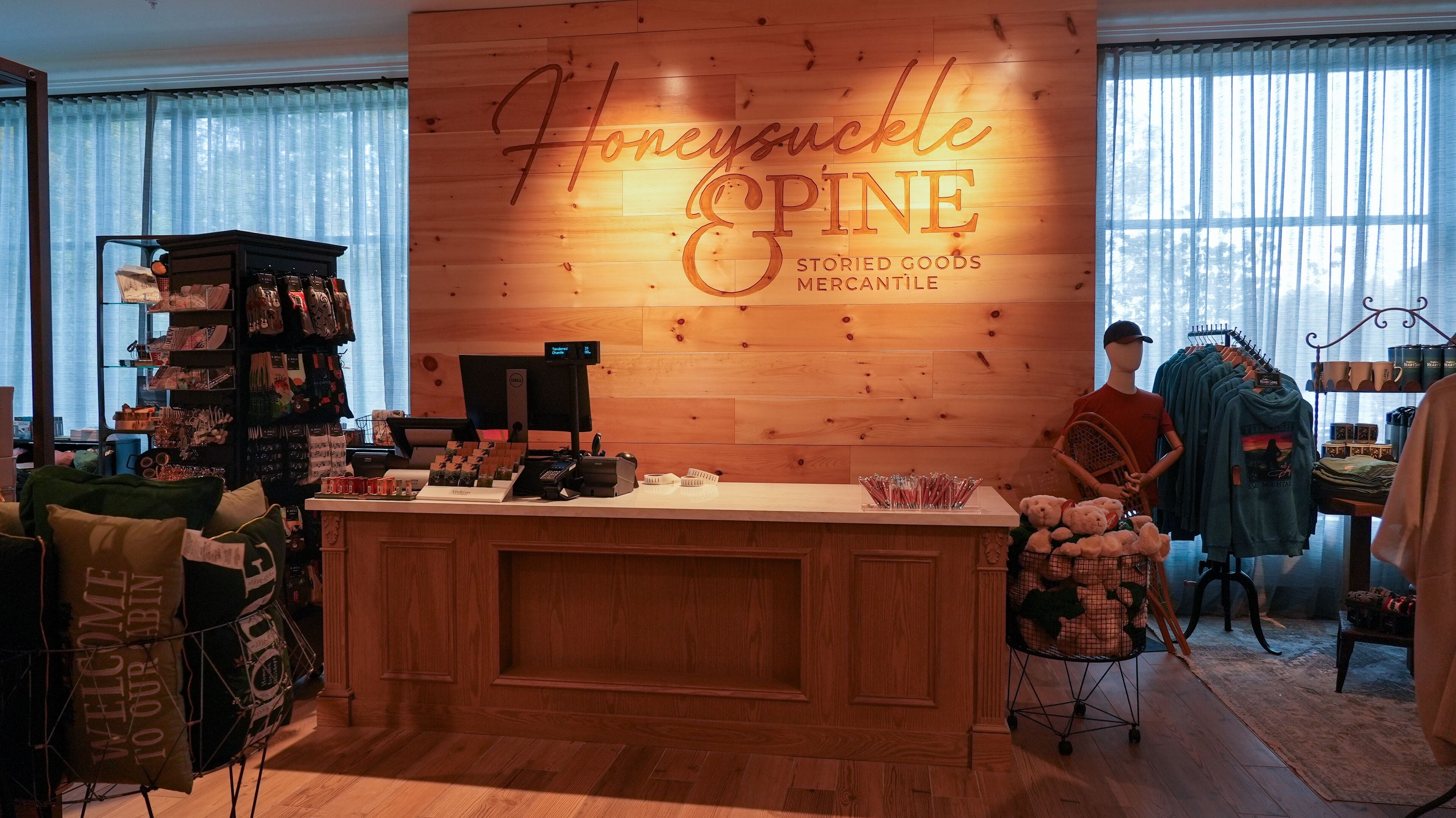 Un acogedor interior de tienda con una pared de madera que muestra 'Honeysuckle Pine' y 'STORIES GOODS MERCANTILE'. Estantes con varios artículos y un maniquí al fondo.