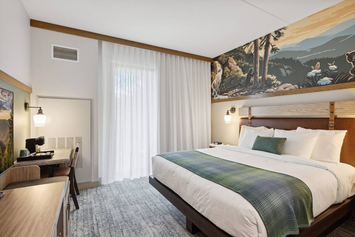 A hotel room with a large bed, a desk with a chair, a painting on the wall, and a window with curtains.