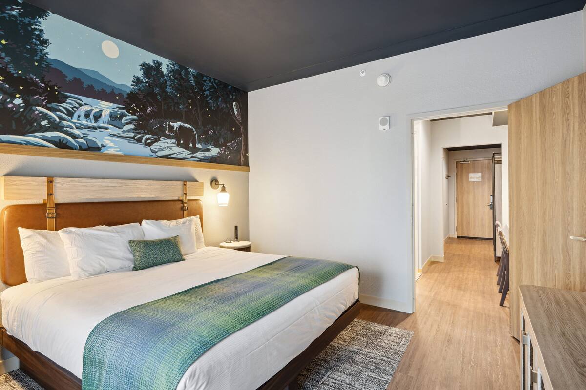 A hotel room with a large bed, a night sky mural on the ceiling, a wooden headboard, and a view into the hallway.