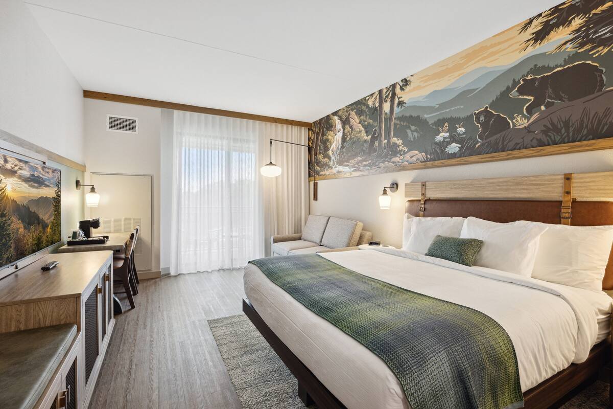 A hotel room with a large bed, a desk, and a painting of a mountain landscape.