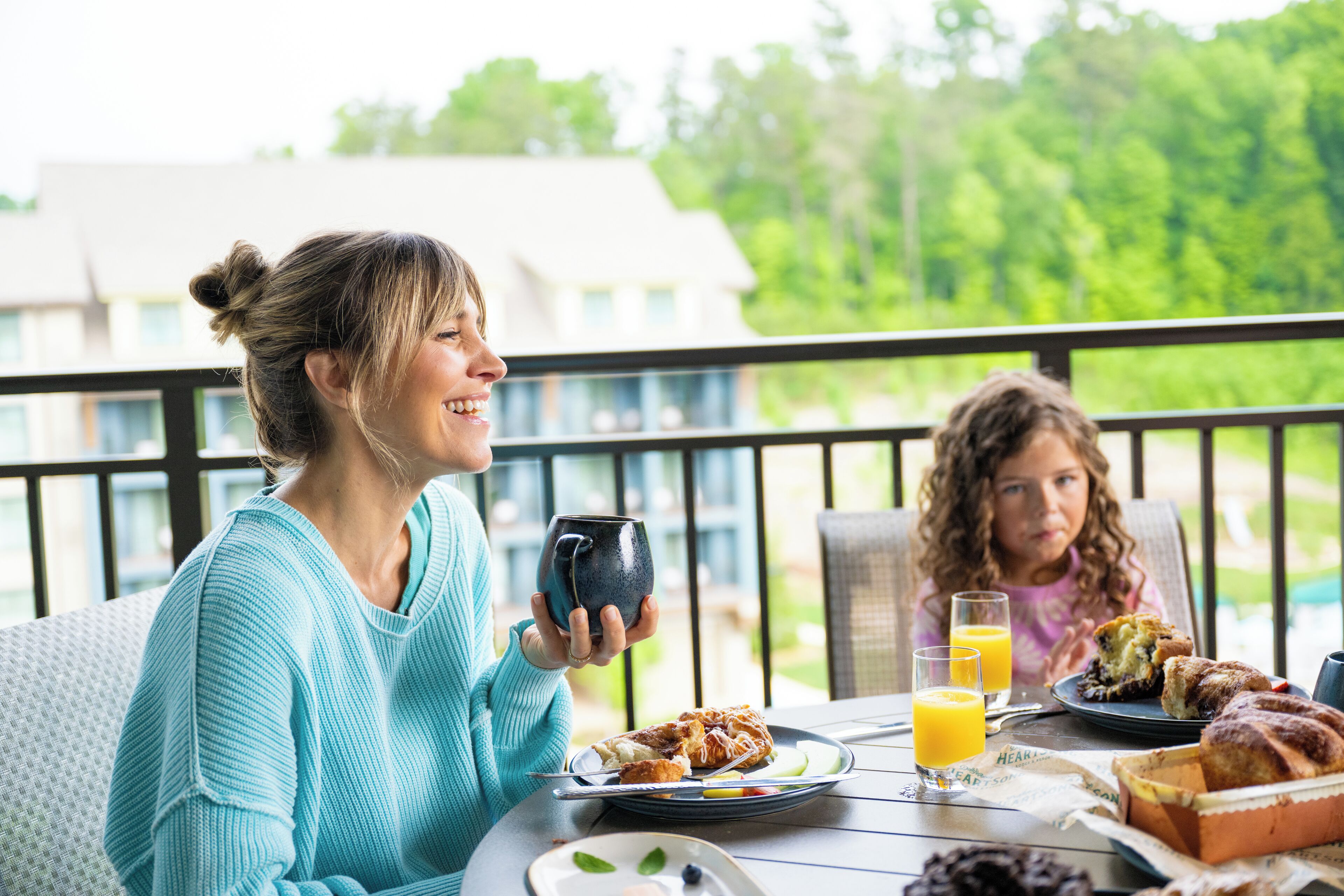 A dining table with food and drinks, a person holding a mug, and a view of greenery in the background.