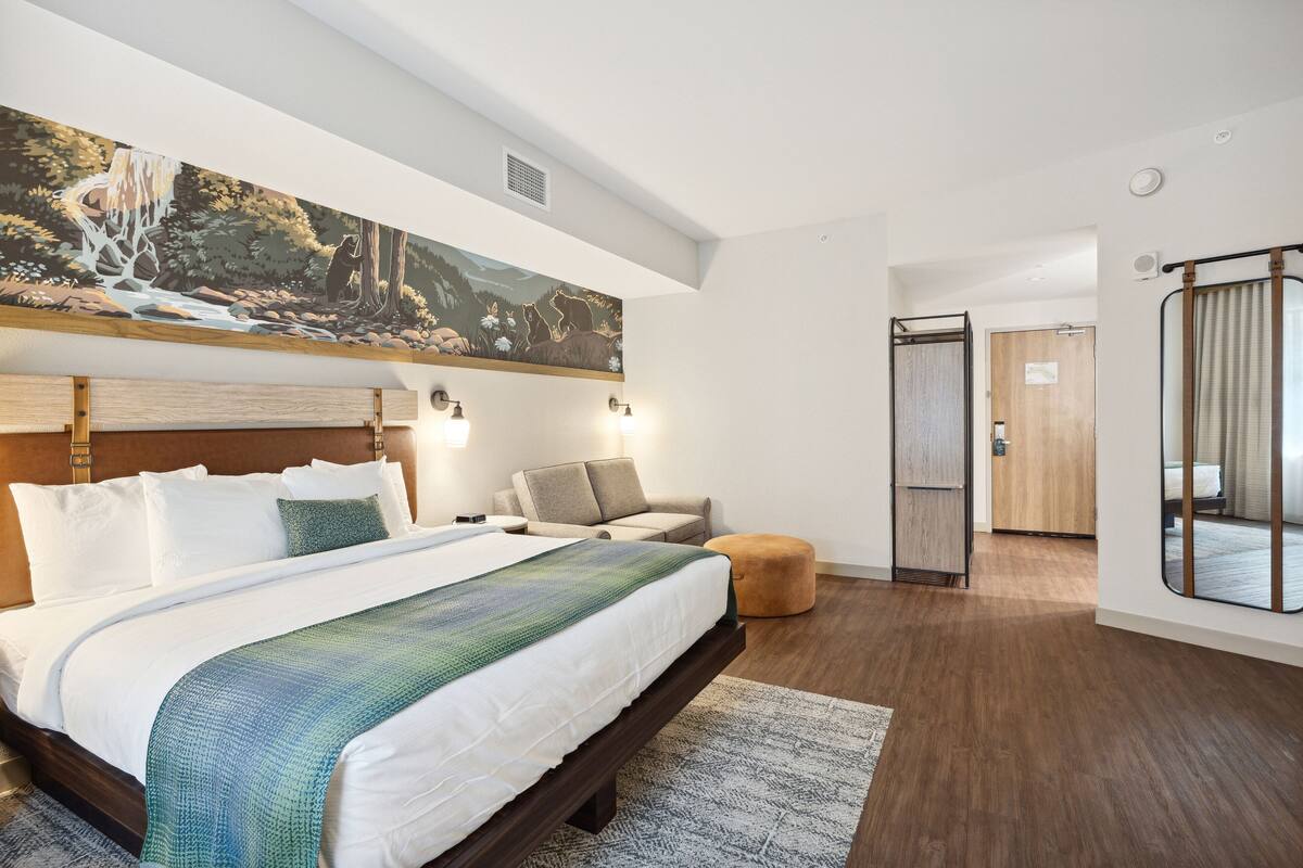 A modern hotel room with a large bed, a seating area with a sofa and armchairs, a wooden floor, and a large painting on the wall.