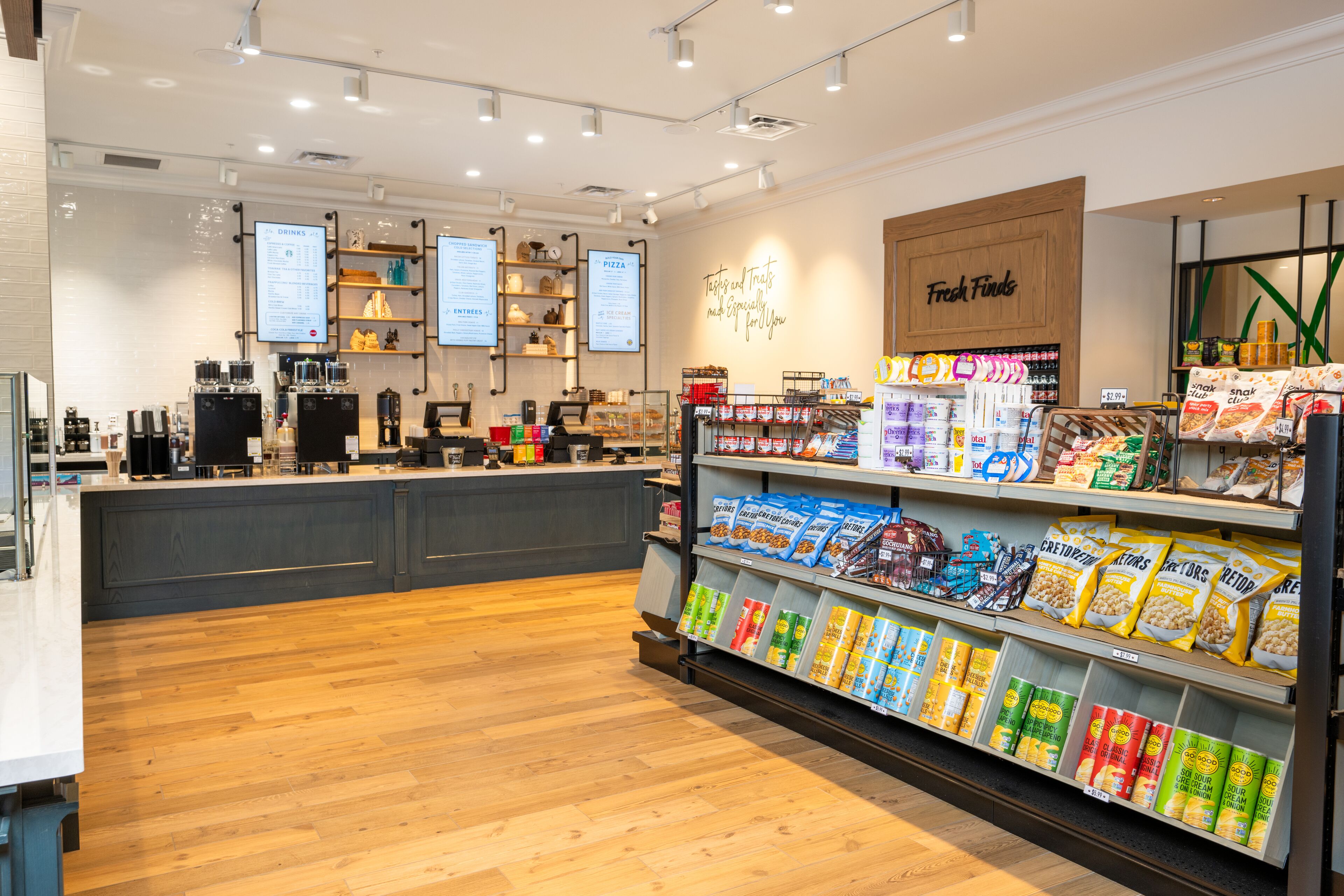 A clean, well-lit store with a wooden floor, a counter with coffee machines, and shelves stocked with snacks and drinks.