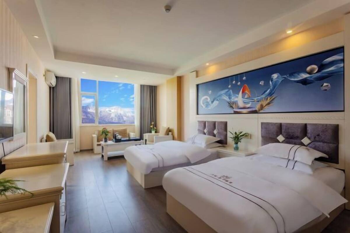 A hotel room with two beds, a desk, a sofa, a painting, and a view of the mountains.