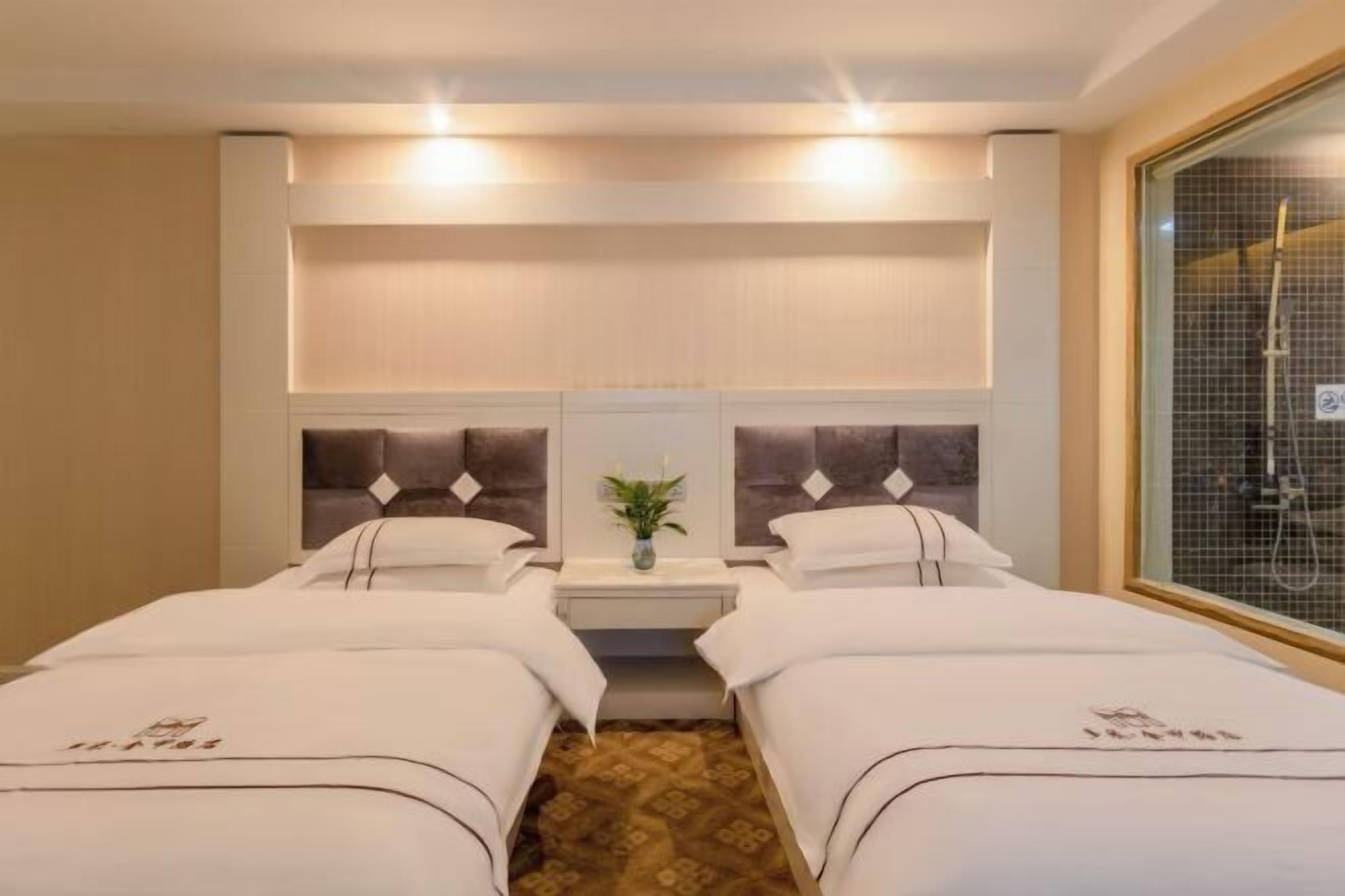 A hotel room with two beds, a small table with a vase, and a window with a shower.