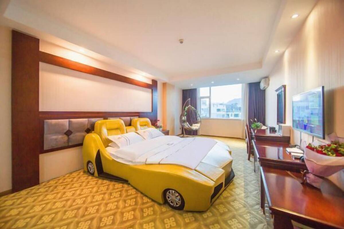 A hotel room with a unique bed shaped like a yellow car, a large window, and a seating area with a TV.