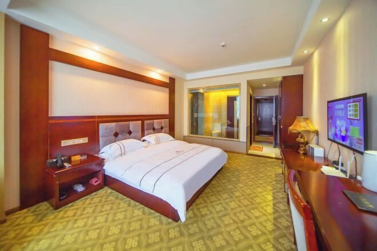 A hotel room with a large bed, a bedside table, a TV, and a desk with a lamp.