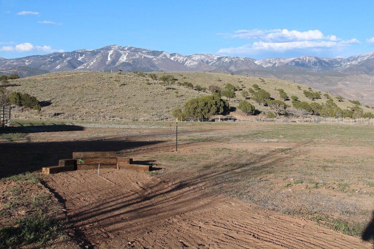 A rural landscape with a dirt path leading to a wooden structure, surrounded by sparse vegetation and distant snow-capped mountains.