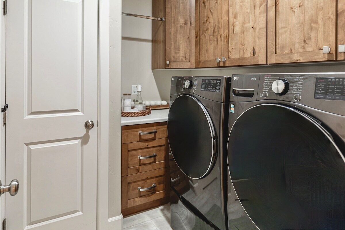A laundry room with two front-loading washing machines, wooden cabinets above, and a white door.
