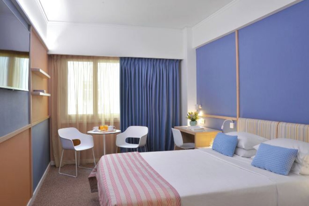 A hotel room with a bed, two chairs, a desk, a small table, and a window with curtains.