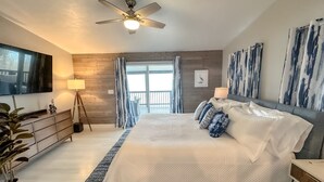 Ein Schlafzimmer mit Bett, Deckenventilator, Fernseher, Kommode, Lampe und einem Fenster mit Vorhängen.