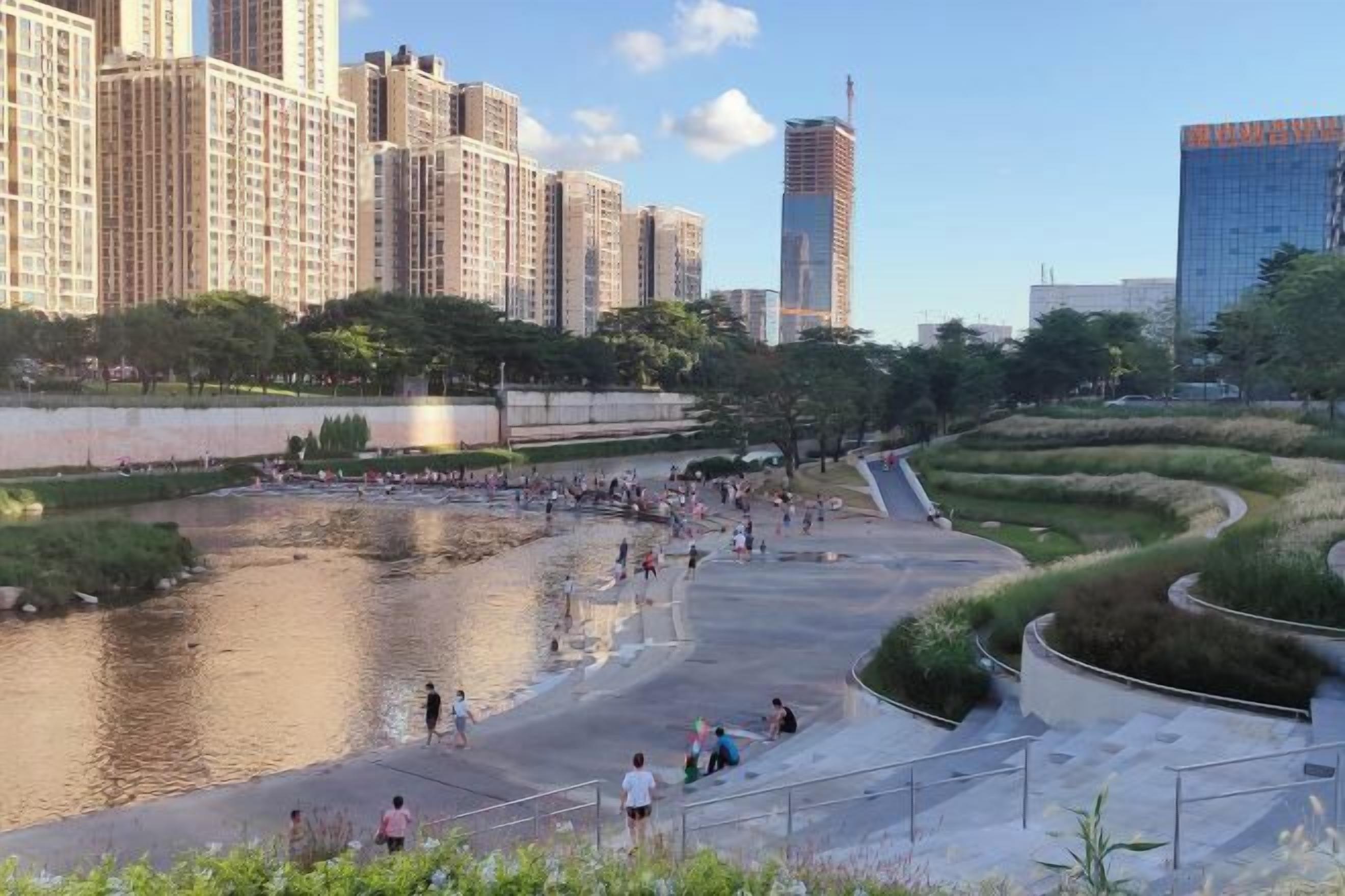 Un parc au bord de la rivière, avec des sentiers pédestres, un pont et de grands immeubles en arrière-plan.
