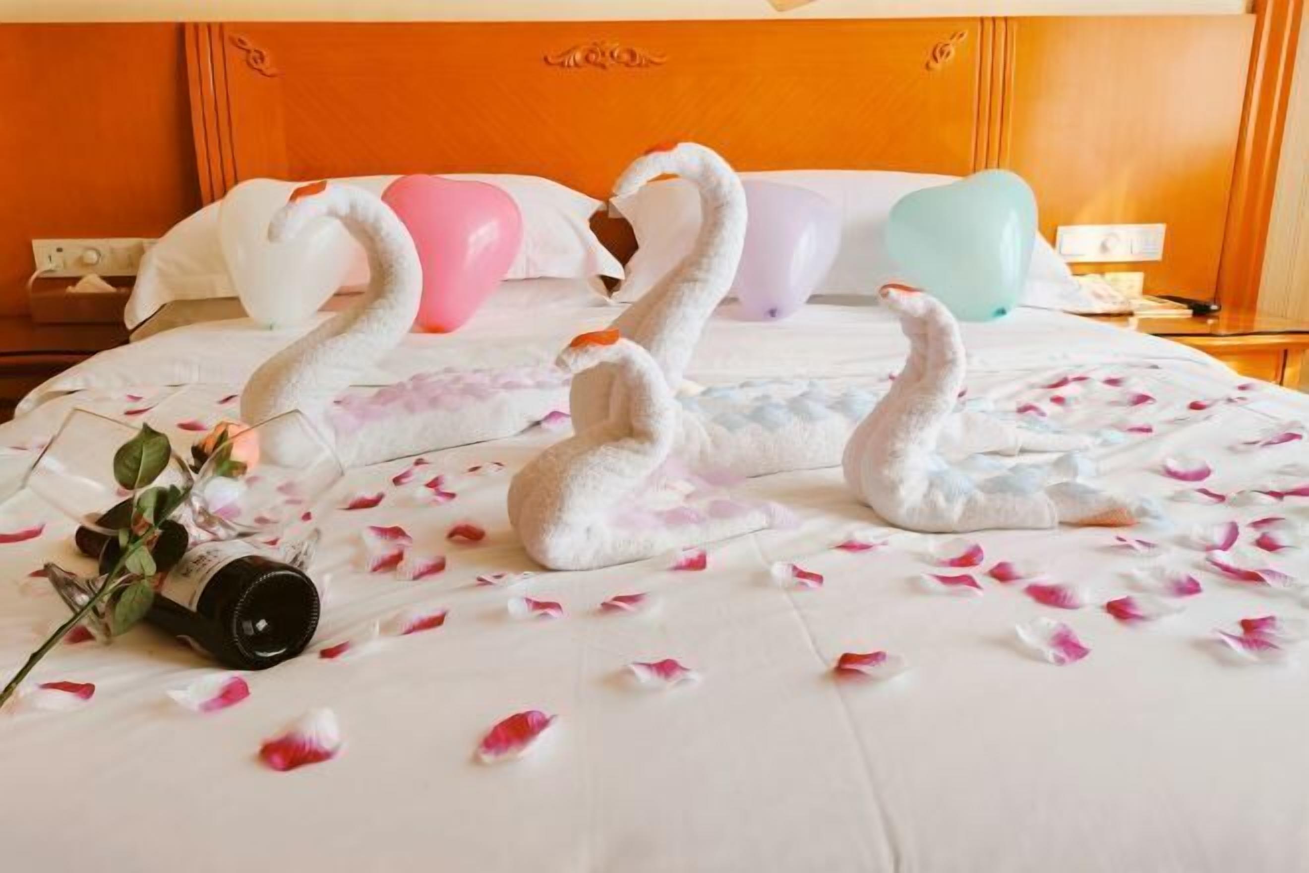 A hotel room with a bed featuring swan-shaped pillows, a bedside table with a lamp, and a decorative vase with flowers.