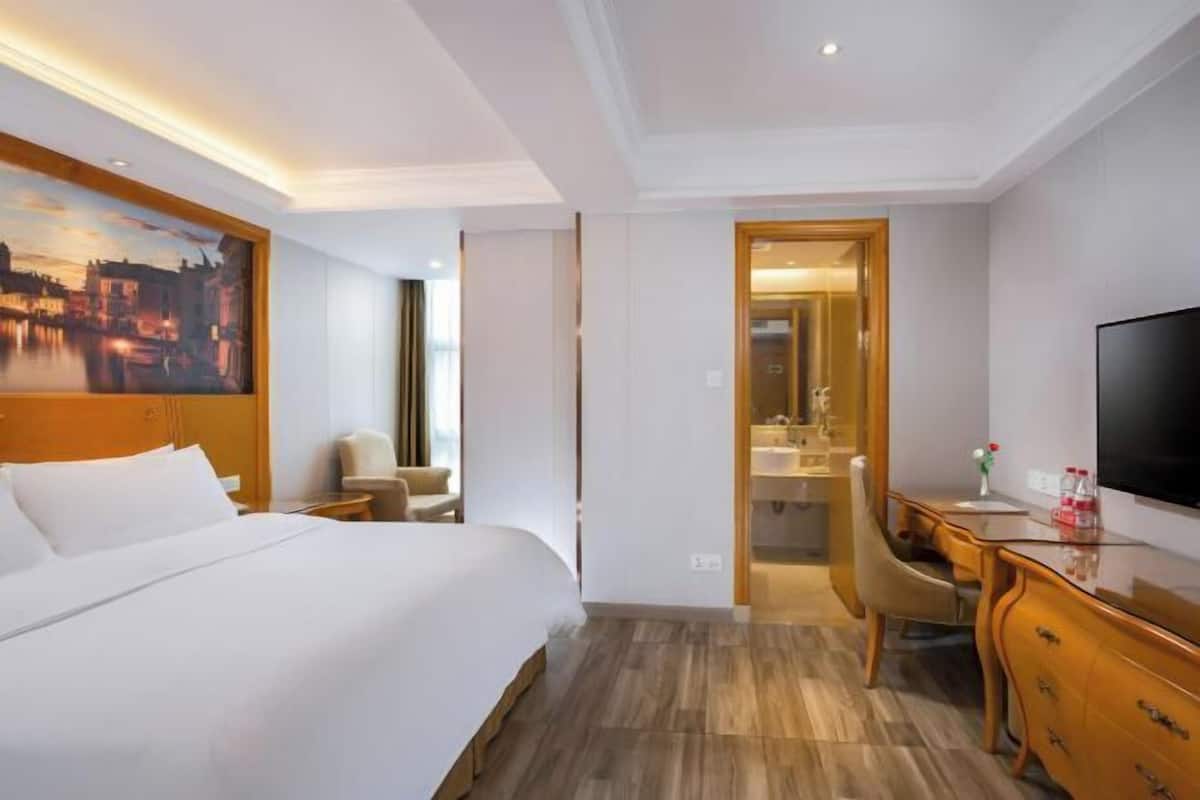 A hotel room with a bed, a desk, a chair, a television, and a painting on the wall.