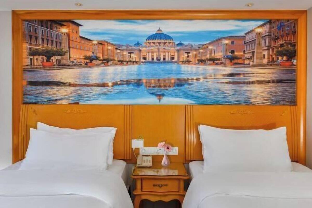 A hotel room with a large painting of a cityscape on the wall, two beds, a nightstand, and a small table with a vase.