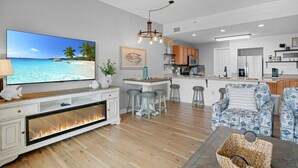 A modern living room with a fireplace, a TV displaying a beach scene, a dining area, and a kitchen in the background. - Luxurious Condo with Zero Entry Pool & Gated Beach (Miramar Beach)