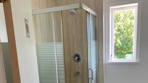 Un espace douche clos par des parois de verre, doté d’un sol carrelé, de toilettes et d’une fenêtre donnant sur un paysage verdoyant.