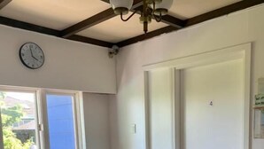Une pièce avec une table en bois, deux chaises, une fenêtre avec des stores, une porte portant un numéro et un ventilateur de plafond.
