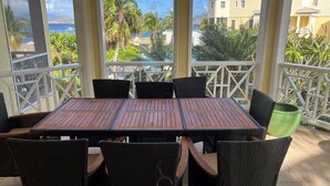 Uma área de estar coberta ao ar livre, com mesa de madeira e cadeiras de vime, com vista para uma praia e edifícios.