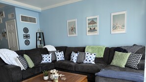 Sala de estar com sofá seccional, mesa de centro e almofadas decorativas. Há decorações nas paredes e uma porta visível.