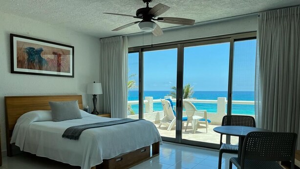 A hotel room with a bed, a ceiling fan, a large window with a beach view, and a dining area with chairs. - Cancun Ocean Front Family Villa 18  at Villas Nizuc (Cancun)