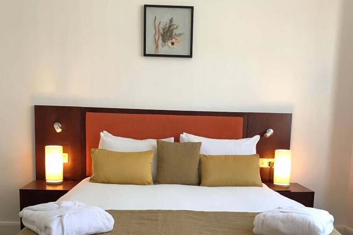 Un lit bien fait, avec une literie blanche et beige, deux lampes de chevet et un tableau encadré au mur.