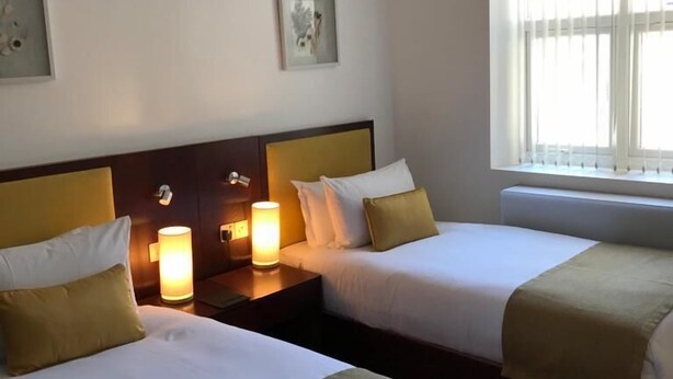 Une chambre d’hôtel avec deux lits, chacun agrémenté de draps blancs et d’un plaid beige. On y trouve deux tables de chevet avec des lampes, et la chambre est dotée d’une grande fenêtre ornée de rideaux légers.