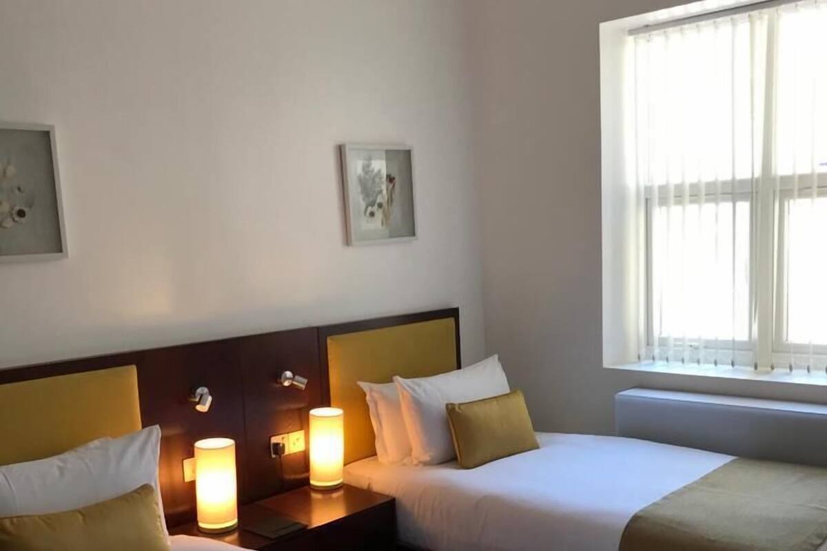 Une chambre d’hôtel avec deux lits, chacun agrémenté de draps blancs et d’un plaid beige. On y trouve deux tables de chevet avec des lampes, et la chambre est dotée d’une grande fenêtre ornée de rideaux légers.