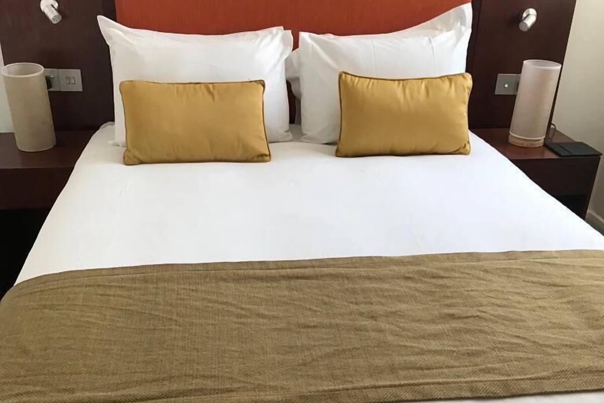 Un lit bien fait, avec des oreillers blancs et moutarde, un plaid marron et une tête de lit en bois.