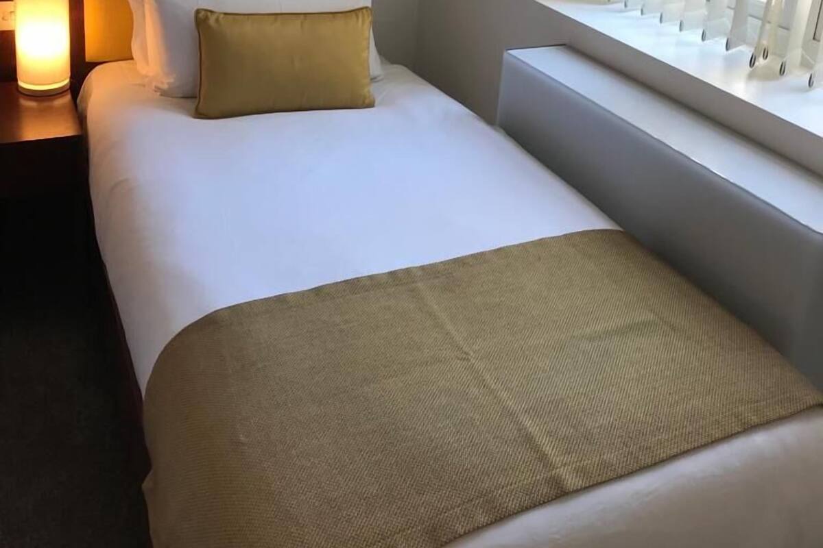Une chambre d’hôtel avec un lit, une table de chevet avec une lampe, une fenêtre avec des stores et un tableau accroché au mur.