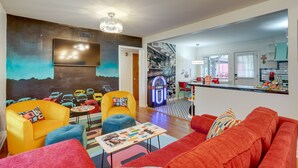 Una sala de estar moderna con mobiliario llamativo, un jukebox retro y una cocina visible a través de un umbral abierto.