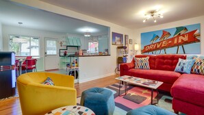 Una sala de estar moderna con un sofá rojo, un sillón amarillo y un reposapiés azul. Se ve una cocina al fondo con un letrero que dice 'Drive-In'.