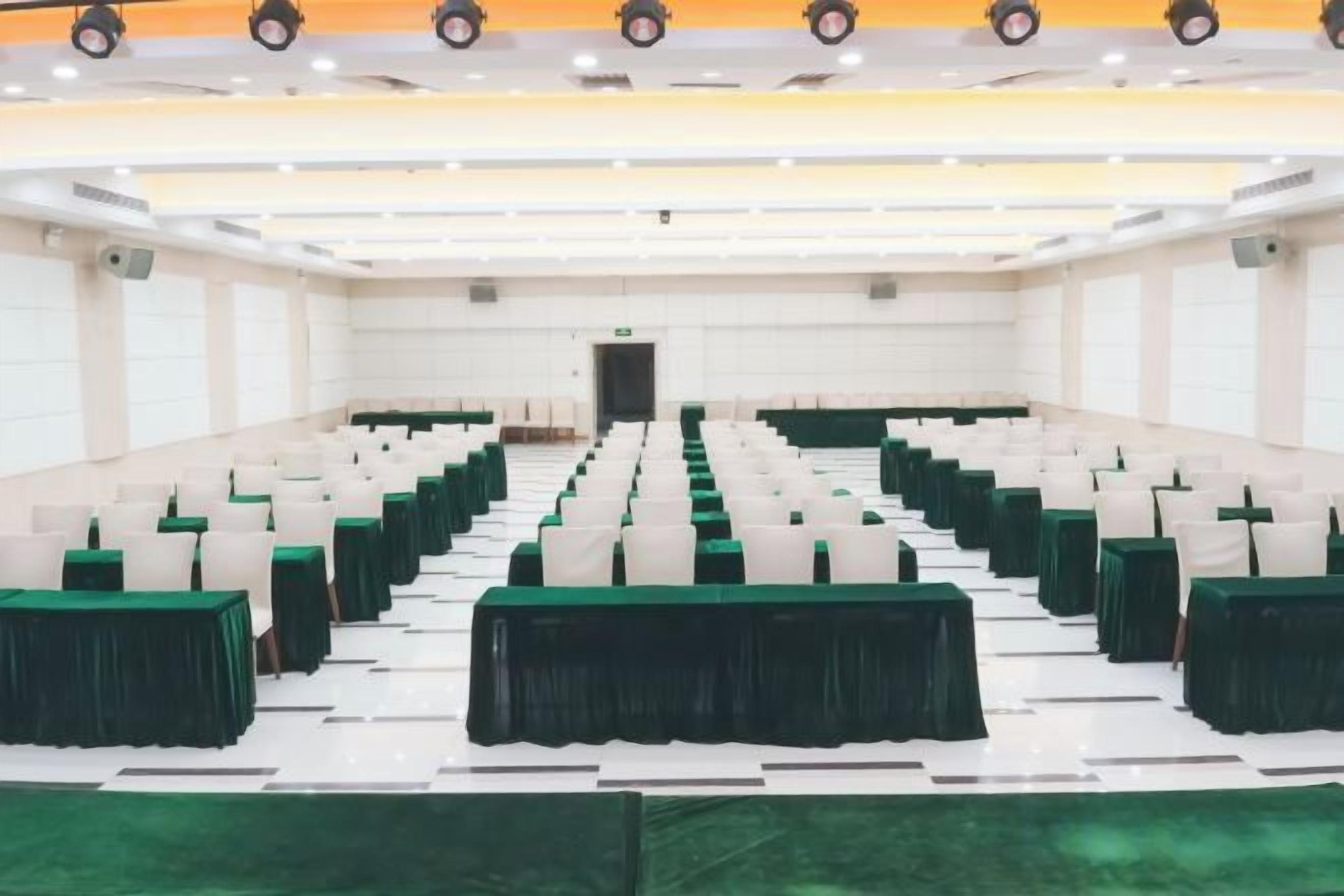 A conference room with rows of tables and chairs, a stage at the front, and a green carpet.