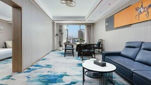 A hotel room with a bed, a sofa, a dining table with chairs, a coffee table, a large window with a city view, and a ceiling light fixture. - Huakang International Hotel (Qianjiang)