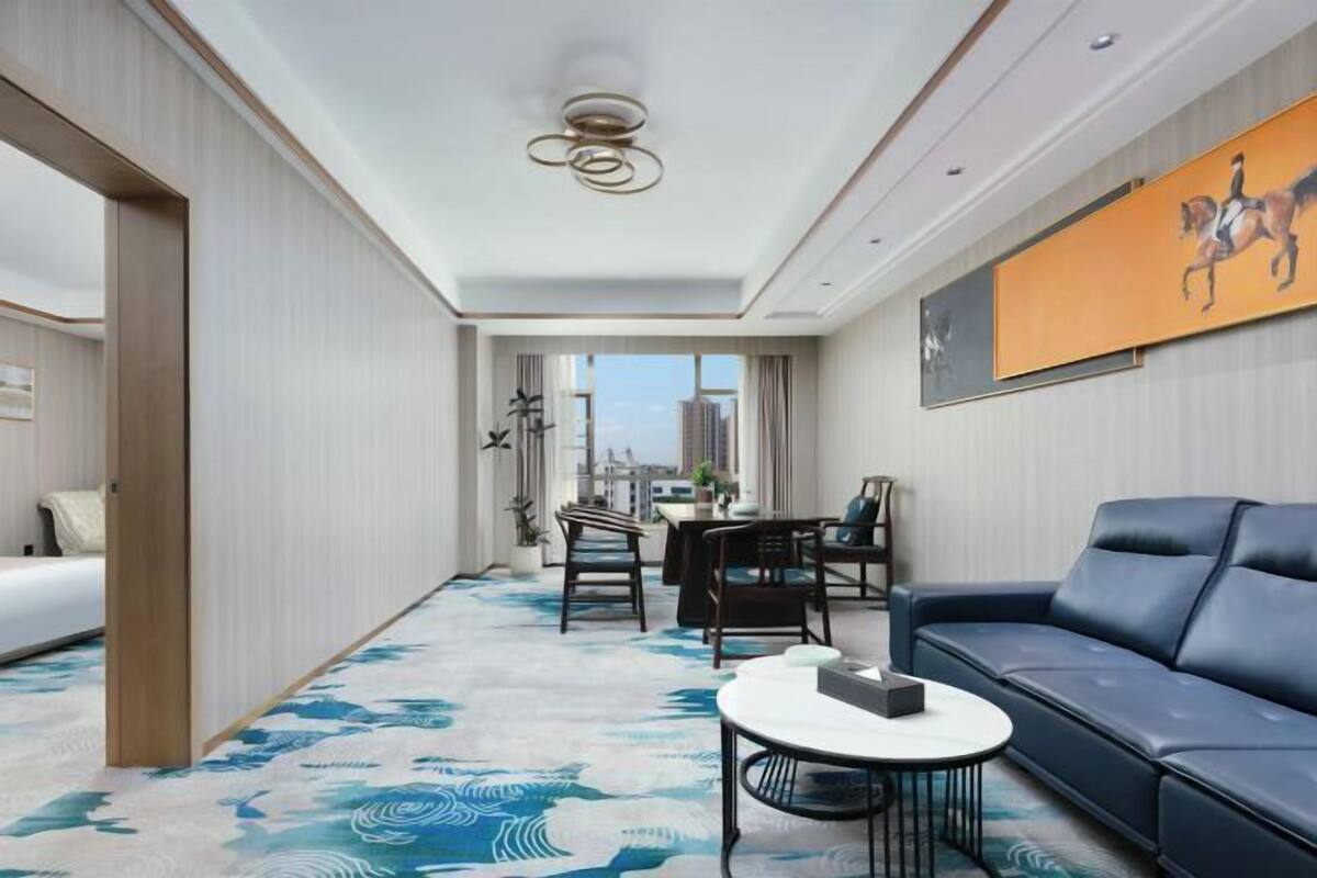 Habitación de hotel con cama, sofá, mesa de comedor con sillas, mesa de centro, ventana grande con vista a la ciudad y lámpara en el techo.