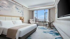 A hotel room with a bed, a desk, a chair, a TV, and a large window with a city view. - Huakang International Hotel (Qianjiang)