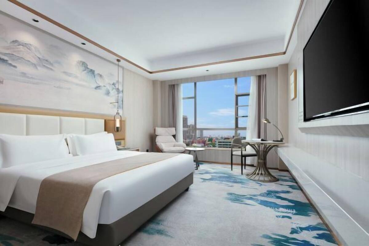 Habitación de hotel con cama, escritorio, silla, televisor y una ventana grande con vista a la ciudad.